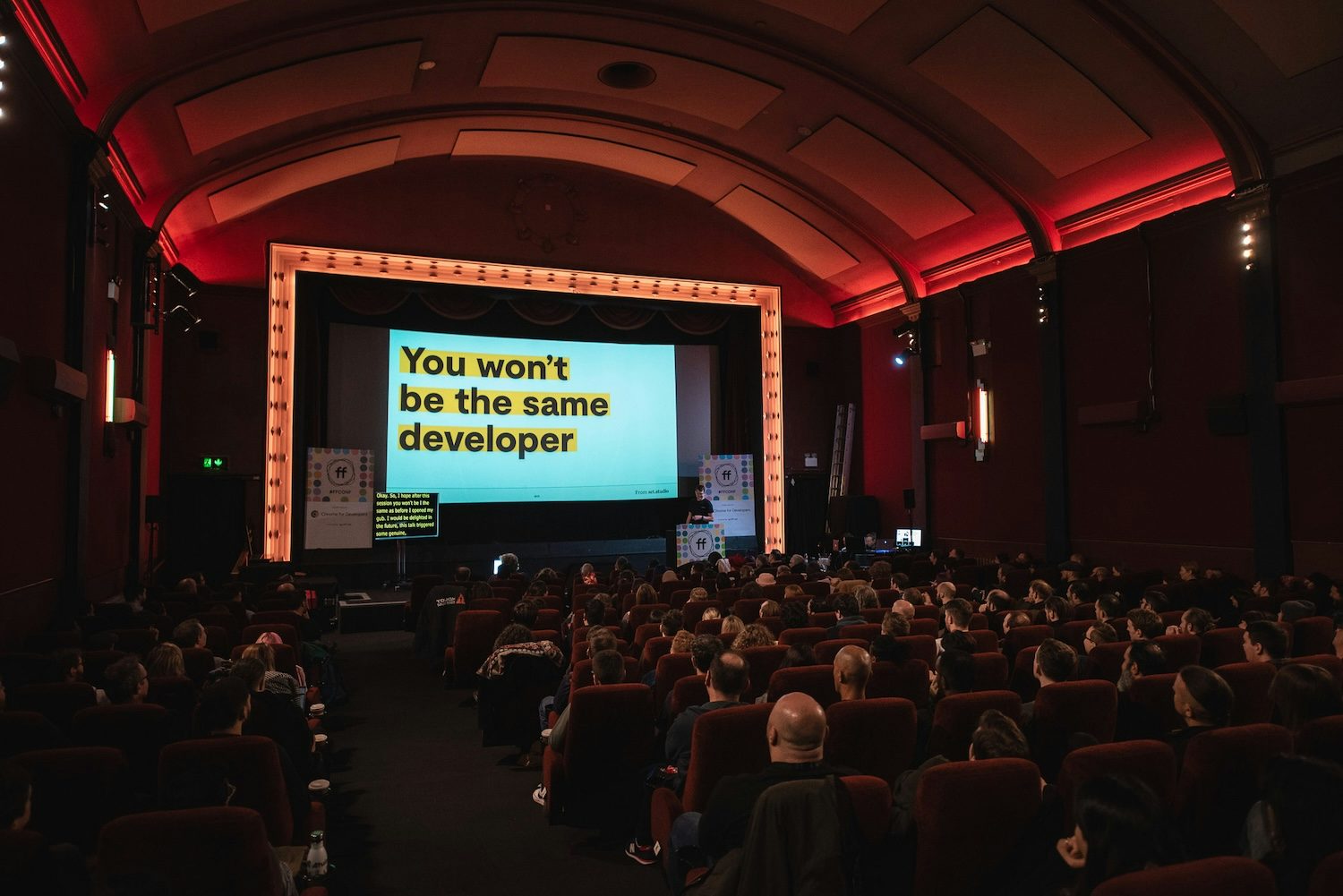 An audience seated in a theater, looking at a large presentation screen with the slide text, 'You won't be the same developer,' in bold yellow text on a black background. The room is dimly lit, with red ambient lighting along the arched ceiling and walls. I'm stood at an FF Conf branded podium to the right of the screen